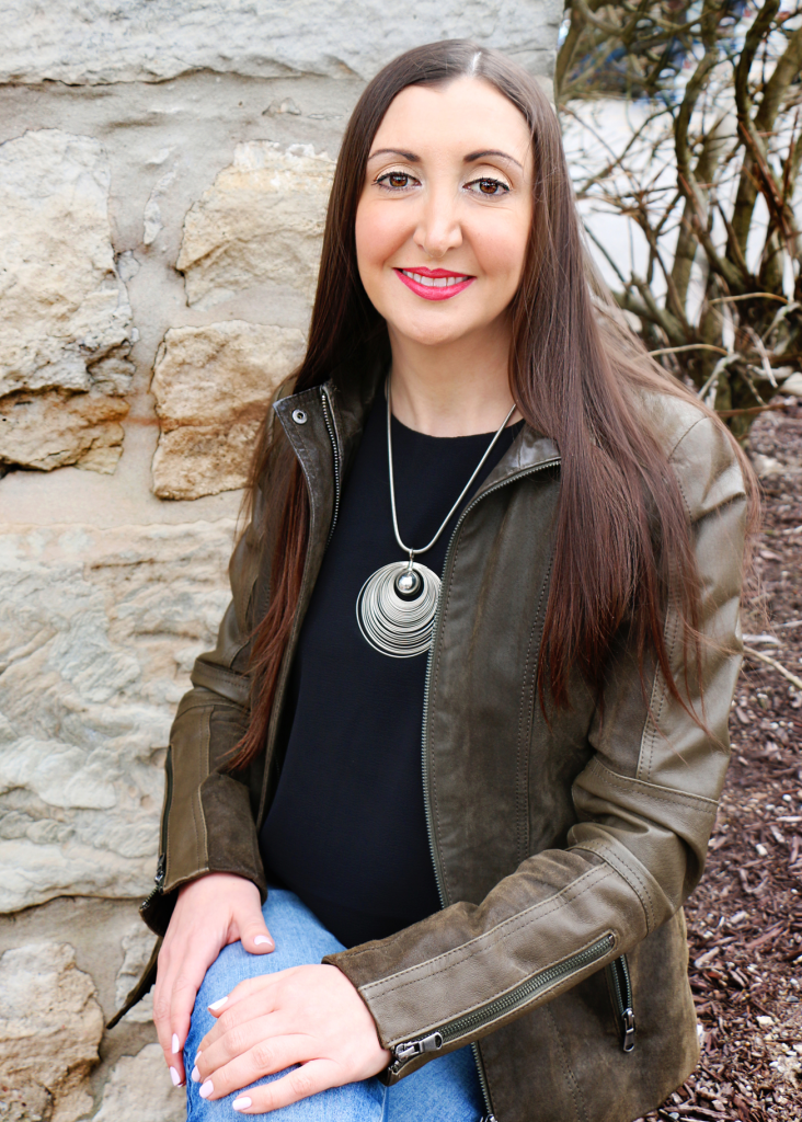 Portrait of author Jade Raven with a stone wall background. Jade has long brown hair, worn down. She is wearing a black shirt, a silver-tone necklace, and a green leather jacket.